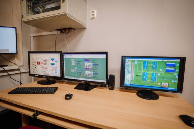 Valmiera, Letonya - 25 Kasım 2024 - Tahta bir masada SCADA sistemleri ve endüstriyel şemaları gösteren üç bilgisayar monitörü.