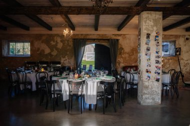 Cesis, Letonya - 6 Eylül 2024 - Beton bir sütun ve doğal ışık üzerine bir fotoğraf kolajının yer aldığı zarif sehpaları, sandalyeleri ve dekorasyonları olan kırsal bir ziyafet salonu.