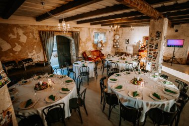 Cesis, Latvia - September 6, 2024 - Elegant event setup with round tables covered in white tablecloths, green napkins, floral centerpieces, and rustic decor in a cozy indoor space. clipart