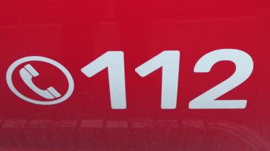 Valmiera, Letonya - 28 Kasım 2024 - Kırmızı arka planda yardım ve kurtarma hizmetlerini sembolize eden 112 numarayı gösteren acil çağrı işaretine yakın plan.