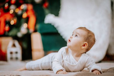 Bebek yumuşak bir battaniyeye uzanmış, merakla yukarıya bakıyor, arkaplanda bulanık Noel dekorasyonu ve parıldayan ışıklarla çevrili..