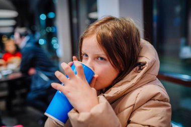 Valmiera, Letonya - 1 Aralık 2024 - Bej ceketli genç bir kız, fast-food restoranındaki büyük mavi bardaktan içiyor, düşünceli bir ifadeyle. Kapalı ortam.