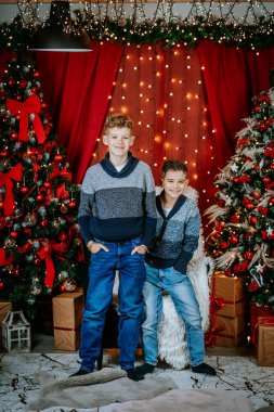 İki kardeş Noel arifesinde ayakta duruyor ve gülümsüyor. Süslü ağaçlar, yanıp sönen ışıklar ve neşeli bir atmosfer yaratan hediyeler..