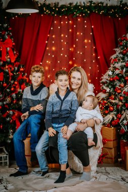 Üç çocuğu olan bir anne, ışıklarla, süslü ağaçlarla çevrili, neşeli bir Noel ortamında poz verir ve hediyeleri rahat bir atmosfere sarar..