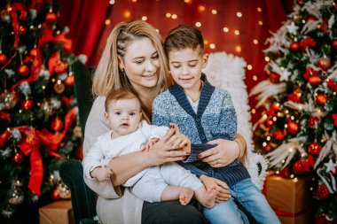 A mother sits with her two children, sharing a moment of joy and connection, surrounded by festive Christmas trees, lights, and decorations. clipart