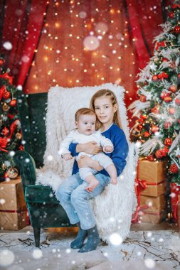 A young girl holds a baby on a cozy chair with festive Christmas decor, lights, and falling snow creating a magical holiday setting. clipart