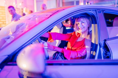 Riga, Letonya - 2 Aralık 2024 - Kırmızı kıyafetli gülümseyen bir kadın Dongfeng arabasında oturuyor, canlı bir ışık ve modern bir atmosferle çevrili..