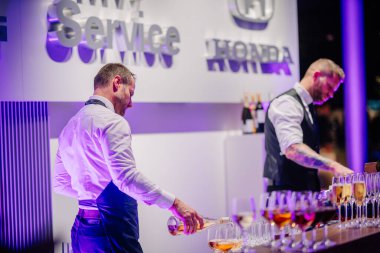 Riga, Latvia - December 2, 2024 - Two bartenders in formal attire serve drinks at a corporate event with a modern bar setup and ambient purple lighting. clipart