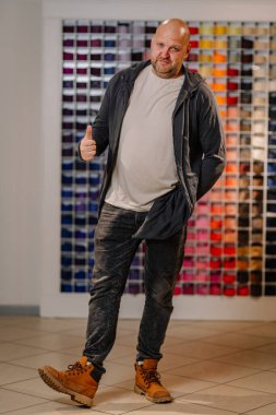 A confident bald man in casual clothing, giving a thumbs-up, standing in a stylish interior with colorful fabric rolls in the background. clipart