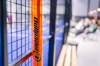 Riga, Letonya - 8 Aralık 2024 - Padel kortunda mavi döşemeli, metal çitli ve arka planda bulanık izleyicili turuncu bir Bullpadel direğinin yakın çekimi. Padel.