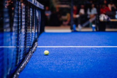 Riga, Letonya - 8 Aralık 2024 - Bir maç sırasında mavi kortta bir padel topunun yakın çekimi. Görüntü, mahkemenin dokusunu ve ağını vurguluyor. Padel.