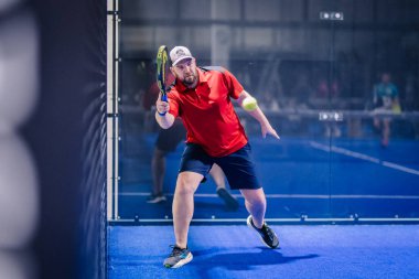 Riga, Letonya - 8 Aralık 2024 - Kırmızı tişörtlü ve mavi şortlu bir erkek padel oyuncusu bir maç sırasında topa vurmak için raketini sallıyor. Padel yarışması