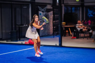 Riga, Letonya - 8 Aralık 2024 - Beyaz elbiseli ve pembe ayakkabılı bir bayan padel oyuncusu kapalı mavi padel kortunda raketiyle topa vurmaya hazırlanıyor. Padel eşleşmesi.