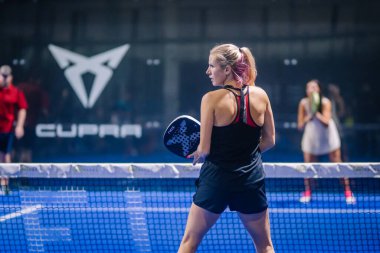 Riga, Latvia - December 8, 2024 - A female padel player holds a racket on a blue court, preparing to serve, with another player visible in the background. Padel match in progress. clipart