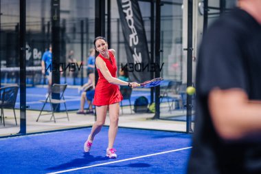 Riga, Letonya - 8 Aralık 2024 - Kırmızı kıyafetli bir bayan padel oyuncusu, kapalı bir mavi padel kortunda raketiyle topa vurmaya hazırlanıyor. Padel oyun eylemi.
