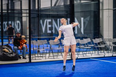 Riga, Letonya - 8 Aralık 2024 - Beyaz spor giyimli bayan bir padel oyuncusu mavi kapalı bir padel kortunda raketle topa vurmak için zıplıyor. Seyirciler ve sandalyeler görülebilir. Padel.