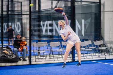 Riga, Letonya - 8 Aralık 2024 - Beyaz spor giyimli bir bayan padel oyuncusu, kapalı alanda mavi bir kortta raketle topa vurmak için zıplıyor. Sandalyeler ve seyirciler görülebilir. Padel.