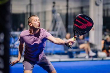 Riga, Letonya - 8 Aralık 2024 - Mor tişörtlü ve gri şortlu bir erkek padel oyuncu kapalı alanda oynanan bir Padel maçında topa raketle vurmaya hazırlanıyor. Padel.