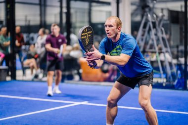 Riga, Letonya - 8 Aralık 2024 - Mavi tişörtlü odaklanmış bir erkek padel oyuncusu, kapalı bir padel kortunda maç sırasında topa vurmaya hazırlanıyor. Padel.