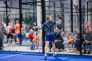 Riga, Letonya - 8 Aralık 2024 - Mavi spor giyim giyen erkek bir padel oyuncusu kapalı bir sahada topa vuruyor ve seyirciler maçı izliyor. Padel.