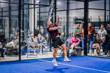 Riga, Letonya - 8 Aralık 2024 - Arka planda seyirci ve oyuncuların olduğu kapalı bir padel sahada oynanan maçta bir erkek padel oyuncusu topa vurmak için atlar. Padel.