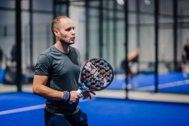 Riga, Letonya - 8 Aralık 2024 - Padel raket tutan erkek bir Padel oyuncusu, mavi bir kortta, kapalı alanda oynanan maçta odaklanmış ve kararlılıkla duruyor. Padel.