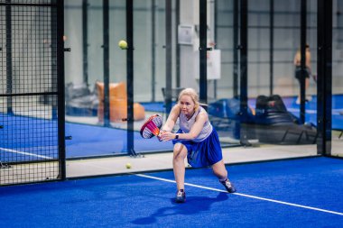 Riga, Letonya - 8 Aralık 2024 - Mavi etekli ve beyaz bluzlu odaklanmış bir bayan padel oyuncusu topu kapalı mavi bir kortta bir padel raketle geri vermek için öne çıkıyor..