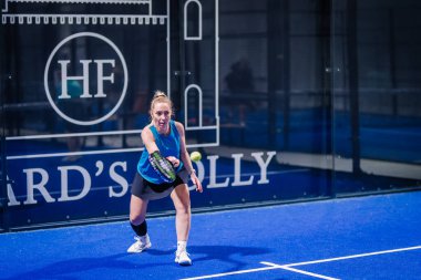 Riga, Letonya - 8 Aralık 2024 - Mavi kıyafetli odaklanmış bir bayan padel oyuncusu, maç sırasında kapalı mavi bir padel kortunda topa vuruyor. Padel spor aksiyon çekimi.