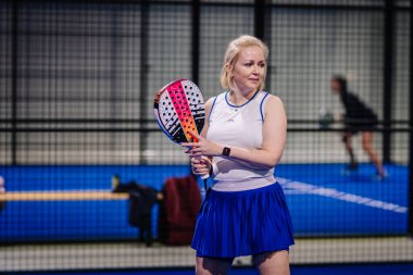 Riga, Letonya - 8 Aralık 2024 - Beyaz ve mavi kıyafetli bir bayan padel oyuncusu mavi bir kapalı sahada oyuna odaklanmış ve maç için hazır. Padel.