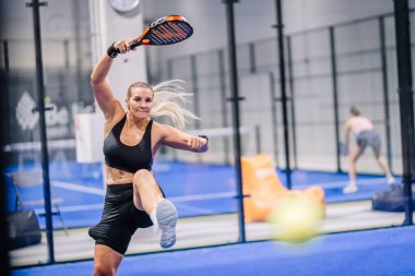 Riga, Letonya - 8 Aralık 2024 - Siyah spor giyimli kadın bir padel oyuncu kapalı sahada bir raketle topa vuruyor, odak ve enerji gösteriyor. Padel.