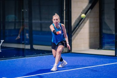 Riga, Letonya - 8 Aralık 2024 - Mavi elbiseli bir bayan padel oyuncusu mavi iç sahada raketiyle topa vurmaya hazırlanıyor. Odaklanmış, dinamik hareket. Padel.