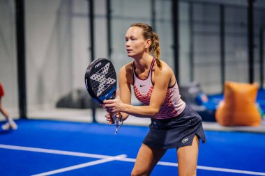 Riga, Letonya - 8 Aralık 2024 - Bir maç sırasında kapalı sahada Padel raket tutan odaklanmış bir bayan Padel oyuncusu. Padel.