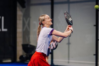 Riga, Letonya - 8 Aralık 2024 - Kırmızı etekli ve beyaz bluzlu bir bayan Padel oyuncusu yoğun bir maç sırasında topa vurmaya hazırlanıyor. Odaklanmış hareket. Padel.
