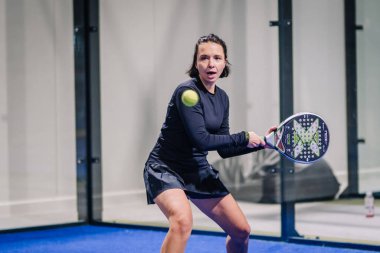 Riga, Letonya - 8 Aralık 2024 - Siyah spor giyimli bayan bir padel oyuncusu kapalı sahada raketiyle topa vurmaya hazırlanıyor. Odaklanmış ifade. Padel eylemi.