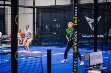 Riga, Letonya - 8 Aralık 2024 - İki bayan oyuncu kapalı bir padel kortunda aktif olarak yarışıyor, raketleri tutuyor ve bir sonraki oyuna hazırlanıyor. Mavi kort ve ağ görünür..