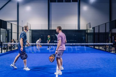 Riga, Letonya - 8 Aralık 2024 - İç saha sahasındaki iki erkek Padel oyuncusu maç sırasında ağların yanında raket tutuyorlar. Mavi kort ve net sahneye hakim..