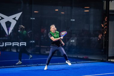 Riga, Letonya - 8 Aralık 2024 - Yeşil tişörtlü bir padel oyuncusu kapalı sahadaki bir maçta raket tutup topa odaklandı. Arka planda Padel markalaşması.