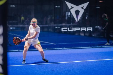 Riga, Letonya - 8 Aralık 2024 - Beyaz spor giyimli odaklı bir padel oyuncusu kapalı bir padel sahada oynanan maçta topa vurmaya hazırlanıyor..