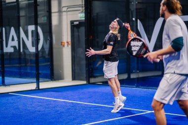 Riga, Letonya - 8 Aralık 2024 - İki erkek oyuncu kapalı bir mavi sahada yoğun bir padel maçında yarışıyor. Bir oyuncu çekim, odak ve çeviklik gösterisi için hazırlanıyor..