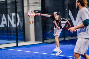 Riga, Letonya - 8 Aralık 2024 - İki erkek oyuncu kapalı bir mavi sahada bir padel maçında yarışıyor. Bir oyuncu atış yapmak için havaya sıçrıyor, gücünü ve dikkatini gösteriyor. Padel eylemi.