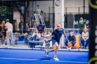 Riga, Letonya - 8 Aralık 2024 - Maç sırasında mavi kortta oynayan erkek bir padel oyuncusu raketiyle topa vurmaya hazırlanıyor. Seyirciler ve oyuncular arka planda. Padel.