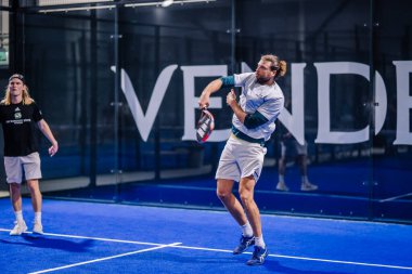 Riga, Letonya - 8 Aralık 2024 - İki oyuncu kapalı bir padel kortunda raket sallayan bir oyuncuyla yarışıyor. Sarayda mavi bir yüzey ve cam duvarlar var..