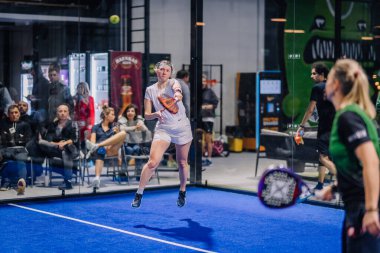 Riga, Letonya - 8 Aralık 2024 - Beyaz giyimli kadın bir padel oyuncusu, mavi sahada oynanan maçta arka planda seyirciler ve başka bir oyuncuyla topa vuruyor. Padel.