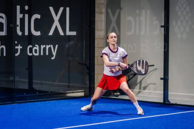 Riga, Letonya - 8 Aralık 2024 - Bir bayan padel oyuncusu kapalı sahada topa vurmaya hazırlanıyor. O mavi zemin ve etrafında cam duvarlar, onun raket tutarak odaklanmış.