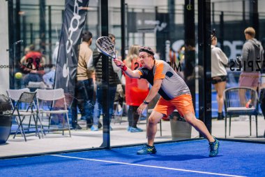 Riga, Letonya - 8 Aralık 2024 - Erkek bir Padel oyuncusu kapalı sahada, raketiyle topa vurmaya hazırlanıyor. Arka planda izleyiciler ve sandalyeler görünür..