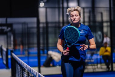 Riga, Letonya - 8 Aralık 2024 - Mavi spor giyimli bir bayan padel oyuncusu ön planda mavi döşeme ve ağ ile kapalı bir padel kortunda raket tutuyor..