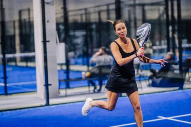 Riga, Letonya - 8 Aralık 2024 - Siyah spor giyimli bir bayan padel oyuncusu mavi kapalı bir padel kortunda raketini sallıyor, odak ve atletik hareketi gösteriyor.