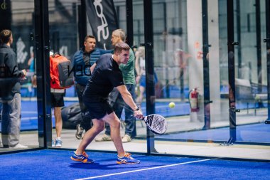 Riga, Letonya - 8 Aralık 2024 - Siyah spor giyimli bir erkek padel oyuncusu arka planda seyircilerle birlikte kapalı mavi bir padel kortunda raketle topa vurmaya hazırlanıyor..
