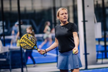 Riga, Letonya - 8 Aralık 2024 - Siyah ve mavi spor giyim giymiş kadın bir padel oyuncusu, kapalı mavi bir padel kortunda harekete hazır sarı raket ve top tutuyor..