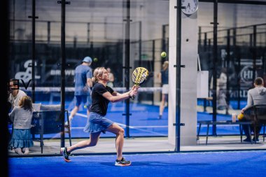 Riga, Letonya - 8 Aralık 2024 - Mavi etekli ve siyah bluzlu bir bayan padel oyuncusu, bir maç sırasında mavi bir padel kortunda sarı raketle topa vurmaya hazırlanıyor..